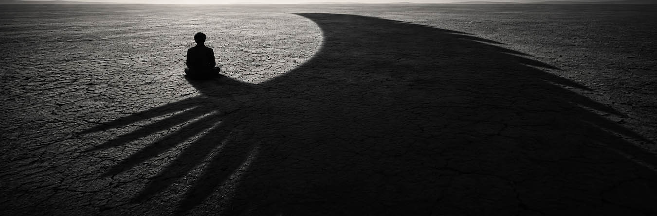 A solitary figure sitting on cracked earth with dramatic shadows — cinematic black and white photograph